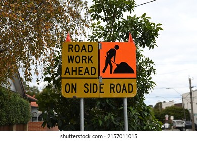 Yellow Road Work Ahead On Side Road Sign, With Orange Square Featuring A Black Icon Of A Man Digging, On A Suburban Street