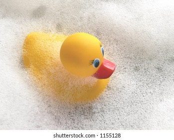 Yellow Rubber Duck With Orange Beak And Blue Eyes Floating In Soap Suds. Overhead Perspective With The Toy Facing From Left To Right. The Soap Suds Are Over The Duck's Back. Copy Space Upper Right.