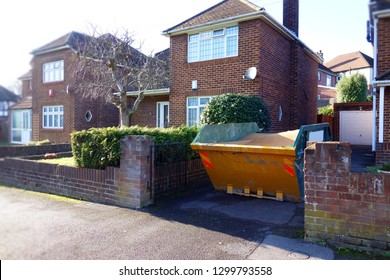 Yellow Rubbish Skip On Driveway. Selective Focus On Metal Skip With Space To Add Text In Front Of The Bin, Footpath, Green Bush Fence, Brick Wall And Houses. Renovate, Moving, Hire Equipment Concept.