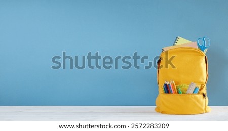 A yellow school backpack with a blue wall behind it. backpack is filled with school supplies such as pencils, pens, and scissors. Back to school organization and school readiness concept