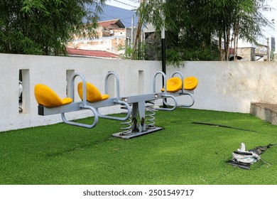 A yellow seesaw sits on a grassy patch in a playground with a white wall in the background. Yellow Seesaw in a Playground Concept Background for Playful Children Activity. - Powered by Shutterstock