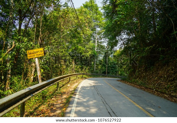 Yellow\
sharp curve warning road sign along local asphalt road through\
natural green forest mountain, Chiangmai,\
Thailand