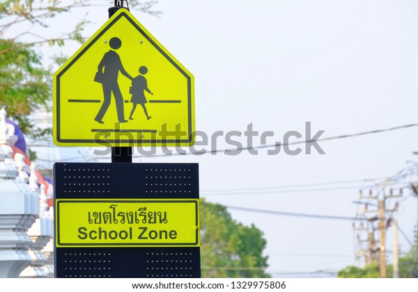 A yellow signboard of School zone on black\
high pole beside the road with white sky background (A Thai\
language on the sign is means School Zone)\
