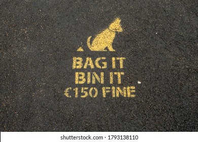 Yellow Sing On A Dark Asphalt To Dog Owners To Clean Up Dog Poo. Bag It, Bin It. 150 Euro Fine.