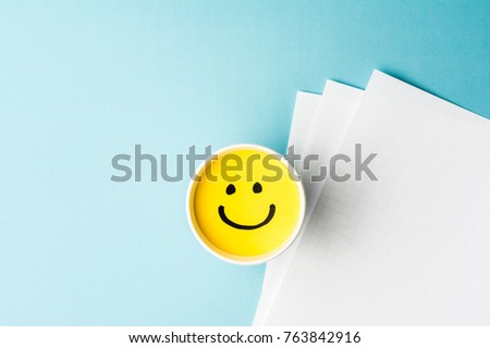 Yellow smiling face, happy mood, on paper cup and papers over blue background.