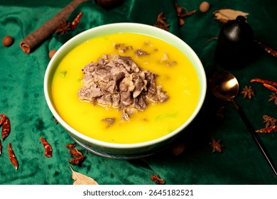 A yellow soup, adorned with sliced beef, is presented in a light green bowl. Star anise and dried chilis are scattered on the dark green fabric. A gold spoon sits beside the dish.