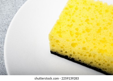 Yellow sponge for dishwashing on a white plate close up on a neutral gray background. Gentle dishwashing. House cleaning. - Shutterstock ID 2259139283