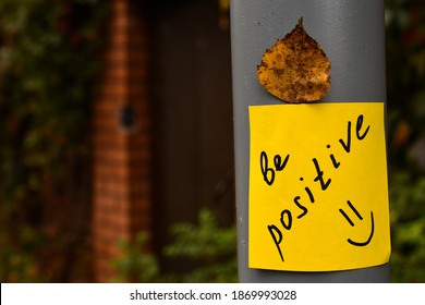 Yellow Sticker With The Words Be Positive And Red Autumn Maple Leaf Glued On A Gray Concrete Pillar Against The Background Of A Red Brick Fence And A Gate