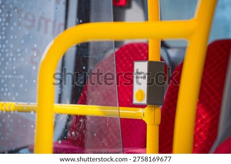 Yellow stop request button inside a public bus with red seats visible in the background and raindrops on the window glass.