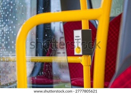 A yellow stop request button on a public bus with red seats and raindrops on the window, indicating a passenger's request to stop the vehicle.