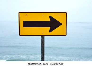 A Yellow Street Sign With Arrow Pointing Right At The Beach With A View Of The Ocean