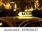 A yellow taxi cab with a lit-up TAXI sign on the roof. The cab is parked in a city at night, with blurry lights in the background.