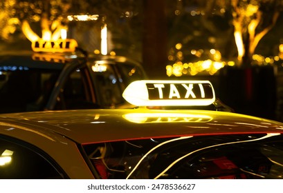 A yellow taxi cab with a lit-up TAXI sign on the roof. The cab is parked in a city at night, with blurry lights in the background.