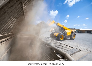 A yellow telescopic loader operates, moving bulk granular material into a ground-level receiving pit at an outdoor industrial or agricultural site, generating substantial airborne dust