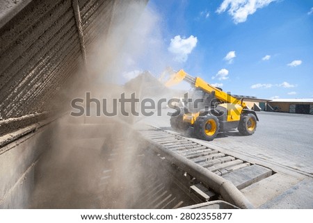 A yellow telescopic loader operates, moving bulk granular material into a ground-level receiving pit at an outdoor industrial or agricultural site, generating substantial airborne dust
