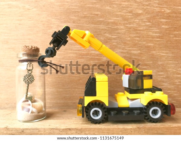 A yellow toy crane is lifting a\
glass bottle inside a bottle of sand and shells on a\
log.