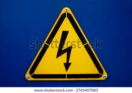 Yellow triangular high voltage warning sign with lightning symbol mounted on blue metal surface in close up.