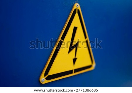 Yellow triangular high voltage warning sign with lightning symbol mounted on blue metal surface in close up.