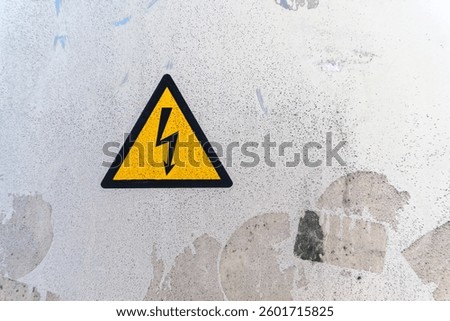 A yellow triangular warning sign with a lightning bolt is prominently displayed on a rough, weathered surface. This signal alerts passersby to an electrical danger.