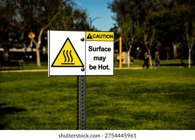 A yellow triangular warning symbol with wavy lines indicates heat, accompanied by text stating 'Surface may be Hot' in a public outdoor space