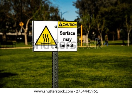 A yellow triangular warning symbol with wavy lines indicates heat, accompanied by text stating 'Surface may be Hot' in a public outdoor space