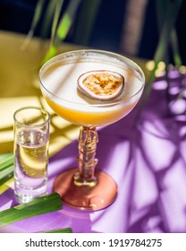 gelber tropischer Cocktail mit Passionsfrucht und Aufnahme auf einem Hintergrund tropischer Pflanzenblätter und Sonnenschirme. Farbhintergrund