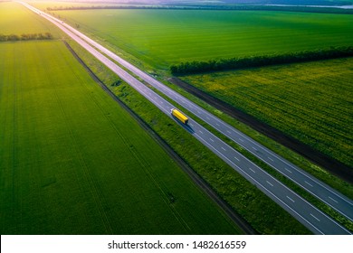 Yellow Truck Driving On Asphalt Road Along The Green Fields. Seen From The Air. Aerial View Landscape. Drone Photography.  Cargo Delivery Left Side Traffic