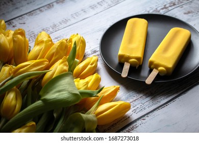 Yellow Tulips And Yellow Ice Cream On A Popsicle Stick Lie On A Light, White Textured Wooden Background. Still Life With Yellow Objects, Illuminated By The Sun. 