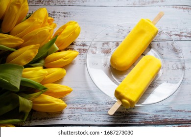 Yellow Tulips And Yellow Ice Cream On A Popsicle Stick Lie On A Light, White Textured Wooden Background. Still Life With Yellow Objects, Illuminated By The Sun. 