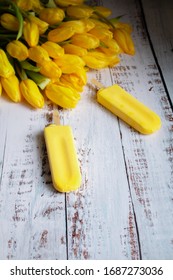 Yellow Tulips And Yellow Ice Cream On A Popsicle Stick Lie On A Light, White Textured Wooden Background. Still Life With Yellow Objects, Illuminated By The Sun. 