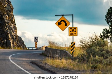 Yellow Warning Road Sign U-turn With 30 Km Per Hour Maximum Speed Posted Under Curve Warning Sign On Roadside, With A Rocky Slope In The Background