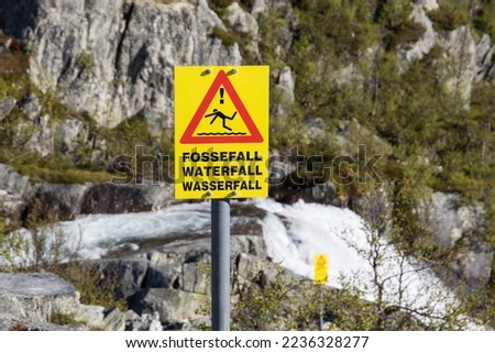 Yellow warning sign in Norway in front of a waterfall with pictorial and bilingual letters Waterfall Wasserfall Fossefall