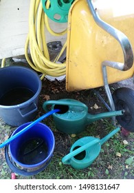 A Yellow Wheelbarrow With A Black Tyre Leaning On A White Wall. A Bucket, A Tub And One Watering Can Are Half Fulled Of Water.In One Pitcher Is A Blue Garden Hose  Filling It Up. Another Tube Hanging.