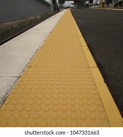 Yellow And White Lines For Public Safety Along The Edge Of An Empty Black Bitumen Train Station Platform Taken From A Low Angle With Forced Perspective