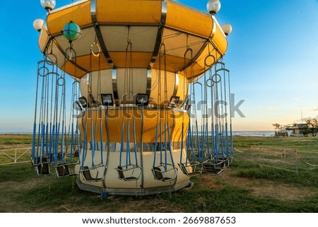 A yellow and white swing carousel with empty seats and blue bars stands still on a grassy area near the coast under a clear blue sky.