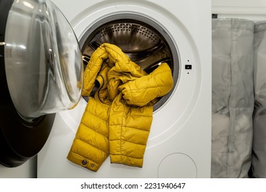 Yellow winter puffer jacket in the drum of open washing machine in laundry room. Washing dirty down jacket in the washer