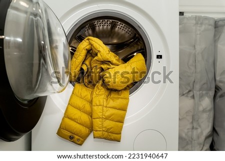 Yellow winter puffer jacket in the drum of open washing machine in laundry room. Washing dirty down jacket in the washer