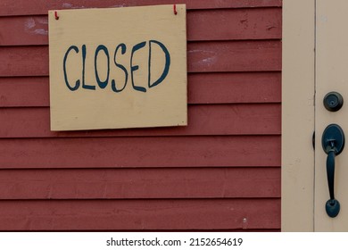 A yellow wooden closed sign with black letters on a red wooden building. The narrow cape cod clapboard wall has a beige colored door with a black metal handle and deadbolt lock. 