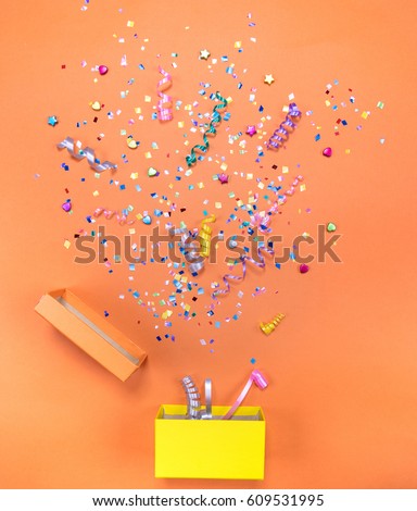 Yelow gift box with various party confetti, streamers, noisemakers and decoration on a orange background. Colorful celebration background.