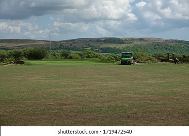 Yelverton England April 2020. Golf Course In Covid-19 Lock Down. View Along Fairway To Putting Green. No Flag, Moorland Beyond. Two Small Work Trucks With Man Repairing  Sand Bunker. Blue Sky, Cloud. 