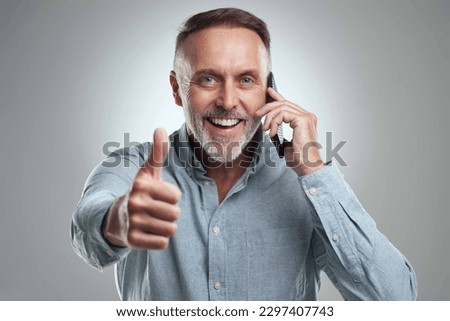 Yep, thats the sound of good news. Studio portrait of a mature man showing thumbs up while talking on a cellphone against a grey background.
