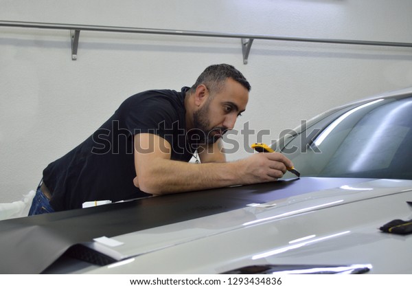 Yerevan, Armenia - 22 January 2019.\
Man\
with beard wrapping vinyls on american car Ford Mustang. Silver\
sport car gonna be with black and red stripes.\
