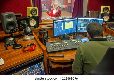 Yerevan, Armenia - December 16, 2020: Operator In Music Studio Control Room And Singer In Booth Behind Glass. Sound Recording Tools, Synthesizer, Audio Mixer And Microphone. Professional Workstation