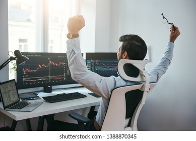 Yes! Back view of stock trader with raised hands looking at multiple computer screens with data and charts and feeling happy while sitting in modern office