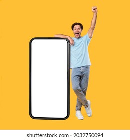Yes, Big Luck. Excited Guy Leaning On Smart Phone With Empty White Screen Shaking Clenched Fist Raising Hand Up, Cheerful Man Celebrating Win, Looking At Camera, Standing At Studio, Mock Up Collage