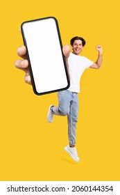 Yes, Cool Mobile Offer Ad. Joyful Young Casual Guy Showing Big Empty Cell Phone Screen Jumping Up In The Air On Orange Studio Background, Creative Collage. Happy Man Recommend Website, Free Copy Space