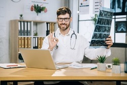 Yes, I Did It. Likable Smiling Male Doctor Scientist, Showing Okay Sign On Camera While Sitting At Desk With X Ray Scan In Hands. Bearded Doctor Using Laptop At Office.