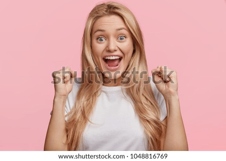 Yes, I done it! Excited cheerful female model with joyful expression, cheers and clenches fists, celebrates her success, has appealing look, poses against pink studio background. Achievement