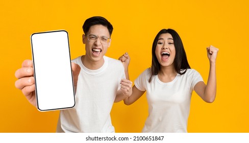 Yes. Excited Asian Couple Holding Big Smartphone With Empty White Cell Screen In Hand Shaking Clenched Fists, Cheerful Guy And Lady Celebrating Win, Yellow Orange Studio Background, Mock Up Copy Space