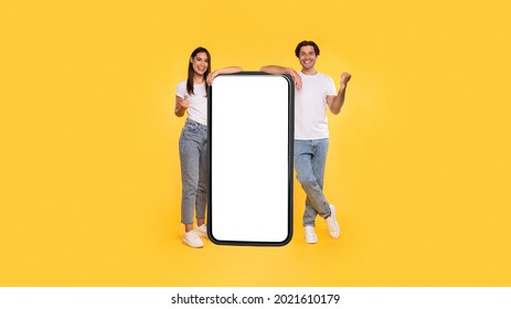 Yes. Excited Couple Leaning On Big Smartphone With Empty White Screen Shaking Clenched Fists, Cheerful Guy And Lady Celebrating Win, Standing On Yellow Background, Mock Up Collage, Full Body Length